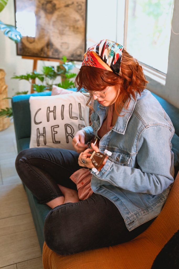Woman with red hair and wearing a jean jacket sitting on a couch playing a ukulele.