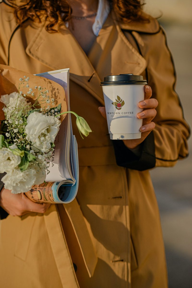Woman in trench coat walking a city street holding a coffee and flowers